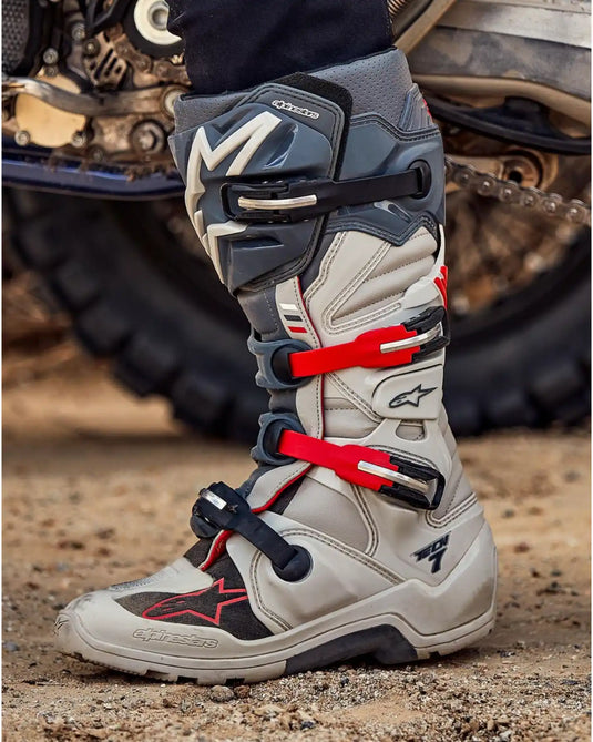 Bota de motocross gris claro y negra con hebillas rojas.