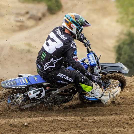 Un piloto de motocross que lleva una camiseta y pantalones negros con acentos blancos y verdes, un casco multicolor y botas blancas, montando una moto de tierra azul y negra en una curva de tierra.